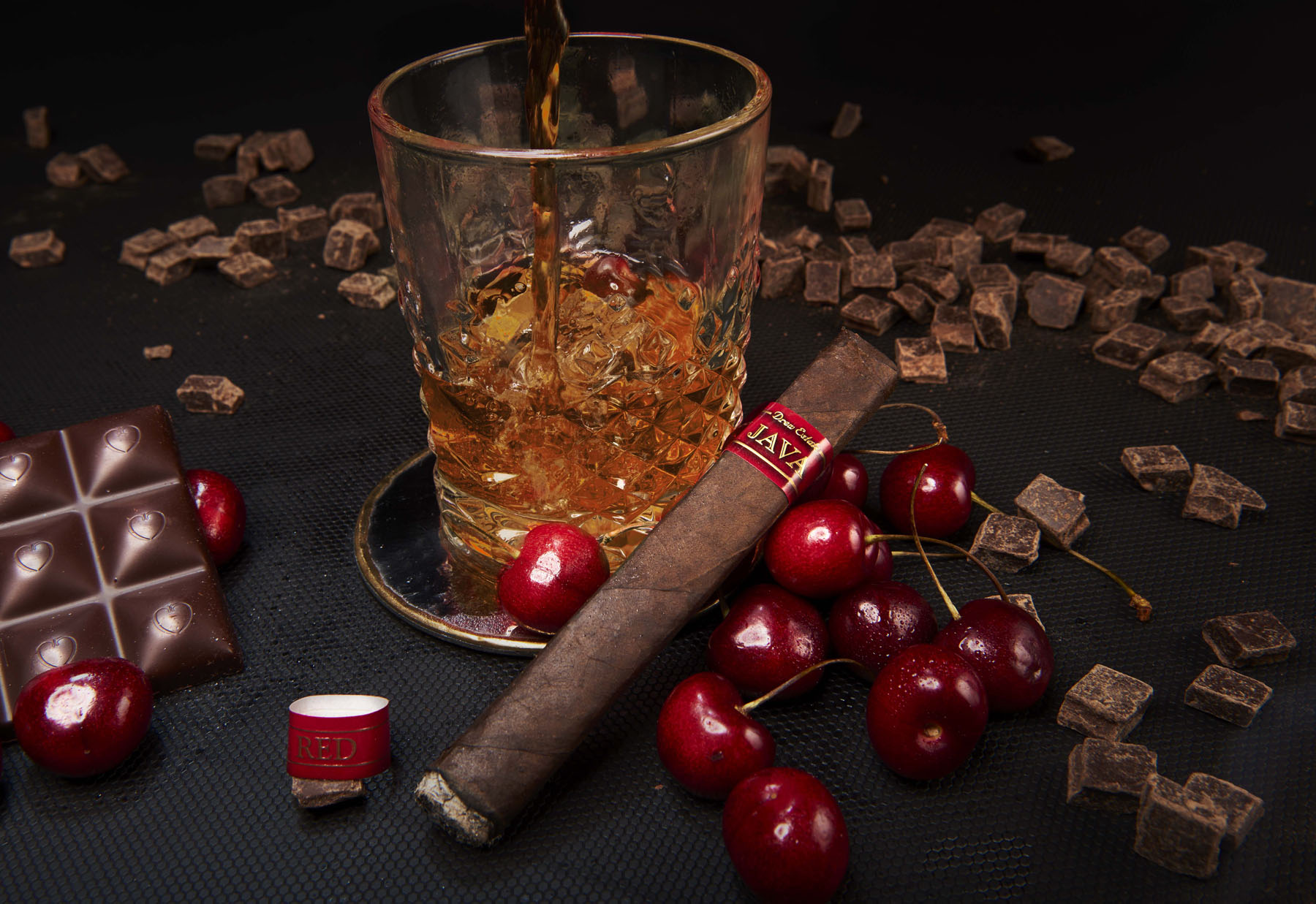 A glass of whisky being poured beside a JAVA cigar, dark chocolate, and ripe cherries on a low table.