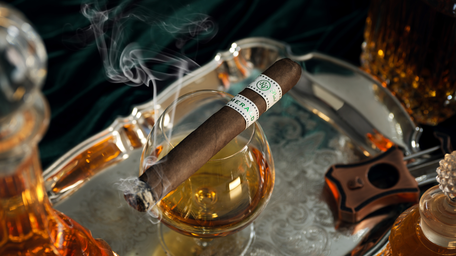A lit cigar resting across a snifter of aged spirit, smoke rising over crystal decanters and a silver service tray.